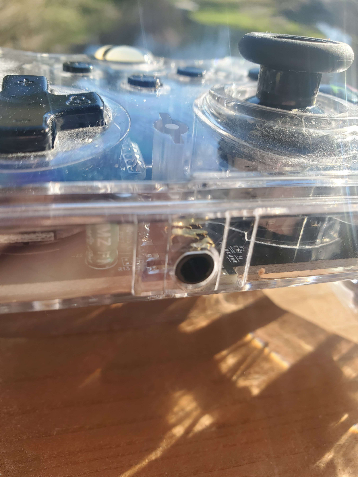 alt-text: pictures of a clear plastic xbox controller. It's shown on my porch-deck with the sunlight streaming in, and there seems to be a sparkle in the sheen of the transparent covering. Perhaps it's reflections from the various circuits and gizmos that are apparent in it's central form, or perhaps it's simply a trick of the light. Upon being placed in shadow, the inner-workings do not appear to sparkle in that same way, so perhaps the beauty is derived from the slivers of the sun that danced across the space between earth and our star. In any case, the entire controller is quite dusty, as if it had been hidden in a paper grocery bag that was shoved in the back of the closet of a boy who doesn't think to clean often. Frankly it's just not worth the trouble, and if he made any messes he would pick them up... but why bother with the little stuff? it's good enough, this is how he lives, so why would we be inconsiderate of his lifestyle? the final picture is of the audio jack.